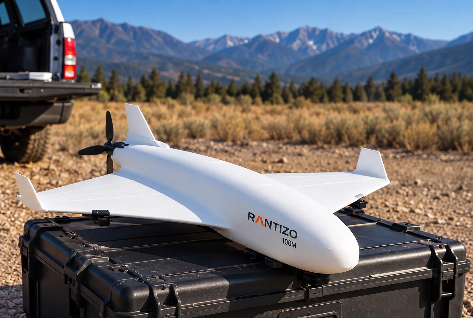 The fixed-wing prototype at rest on its hard-shell carry case, propeller still, a pickup and pine-covered mountains behind